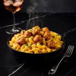 A black bowl filled with creamy garlic mac and cheese, topped with sticky honey garlic chicken cubes, garnished with parsley and black pepper, served on a black marble table with a glass of rosé wine in the background.